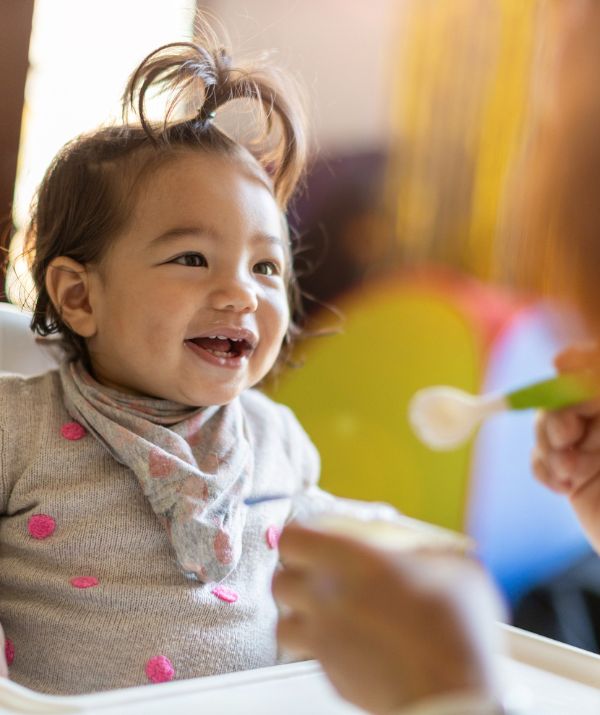 Smiley little girl with a top know pony tail sits in highchair waiting for spoon of yoghurt