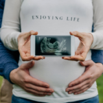 Woman in long white sleeves, holding a baby's ultrasound photo with a man's hand holding her tummy