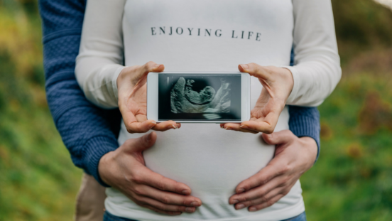 Woman in long white sleeves, holding a baby's ultrasound photo with a man's hand holding her tummy