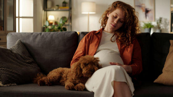 Woman with long red hair, 19 weeks pregnant, sitting on sofa with dog cuddled up