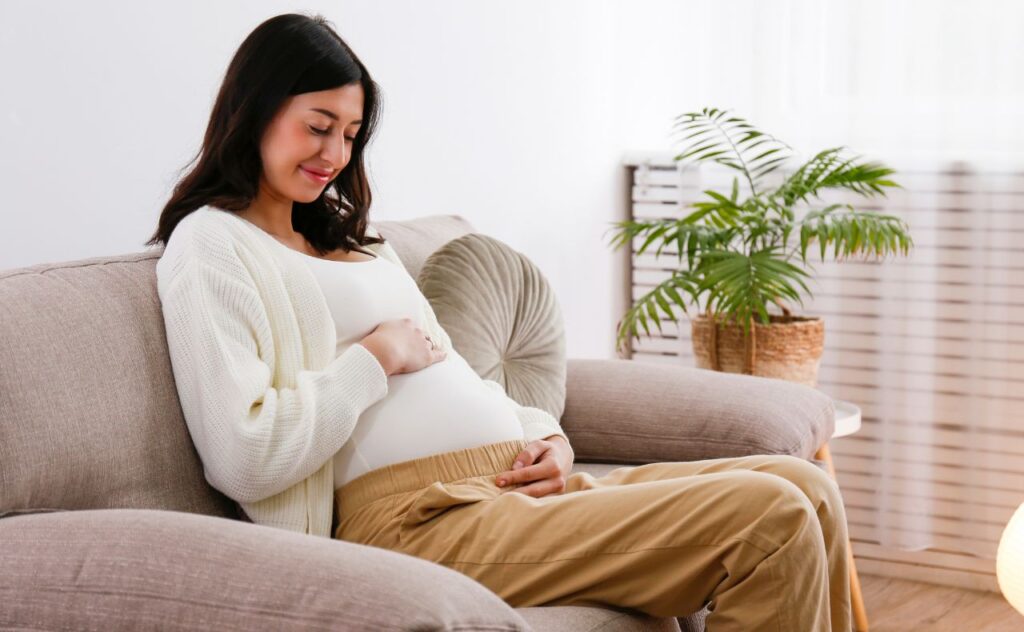 Pregnant woman in second trimester of pregnancy sitting on the couch wearing a white top and white cardigan