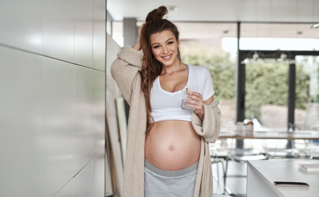 Pregnant woman in kitchen, showing off bare bump, wearing a cardigan and having a drink