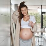 Pregnant woman in kitchen, showing off bare bump, wearing a cardigan and having a drink