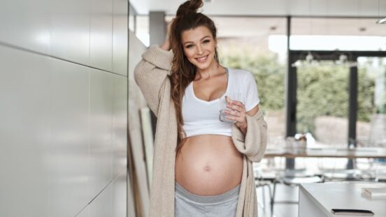 Pregnant woman in kitchen, showing off bare bump, wearing a cardigan and having a drink