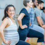 A young and pregnant asian woman has her hands on her hips and stretches her legs, at the front of a line of people doing the same thing.