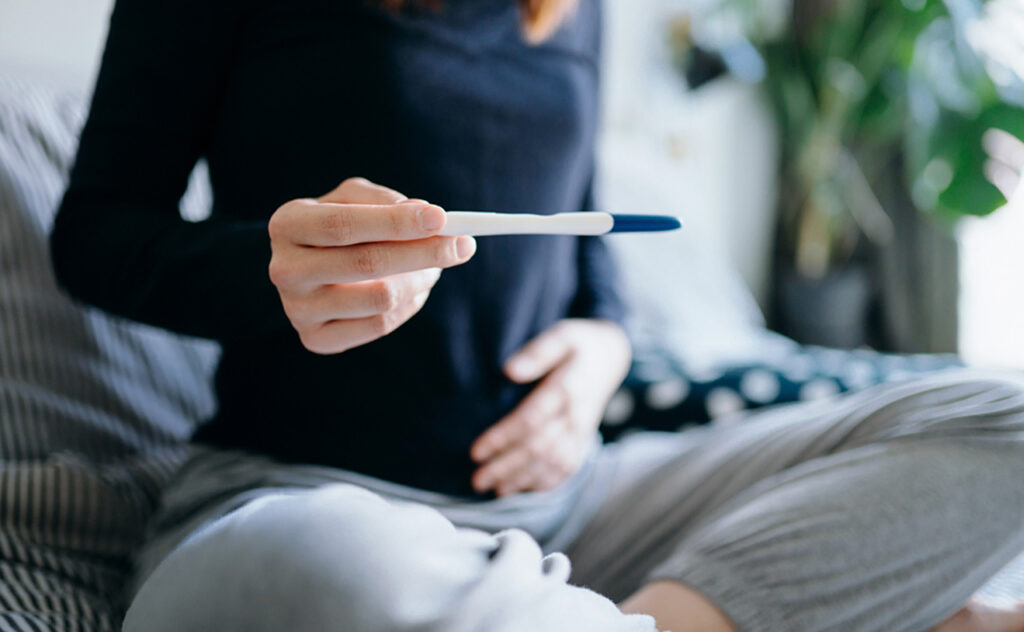 Close up of young Asian woman sitting on the bed holding a positive pregnancy test and touching her baby pump. It's finally happening. 