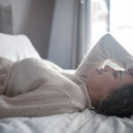 Beautiful young pregnant woman is touching pregnant belly while lying on the bed in bedroom and enjoying morning lights