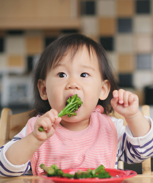 Asian toddler sitting at the table about to take a big bite of broccolini