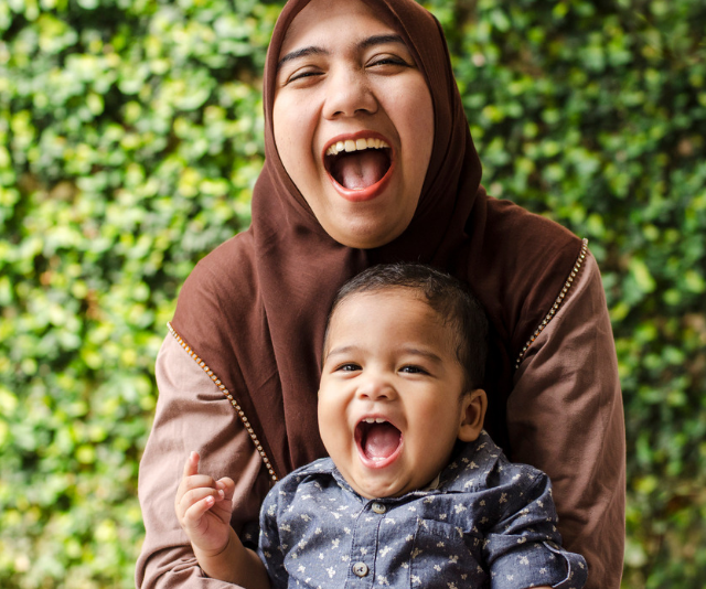 Smiling woman wearing hijab nursing an adorable smiling infant boy with a green foliage wall behind them.