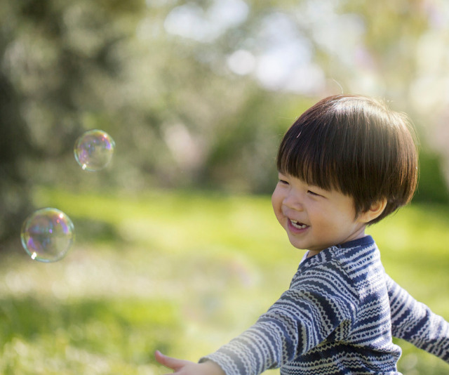Smiling dark haired infant chasing bubbles in a meadow.