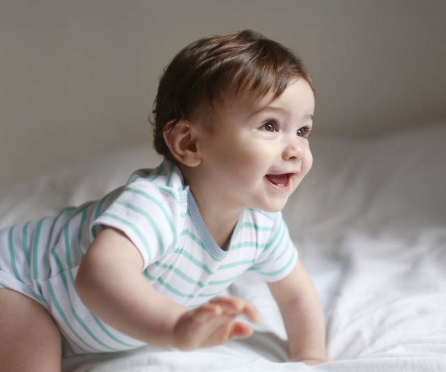Cheeky baby in a blue and white striped onesie crawling across a bed in a sun-filled room.