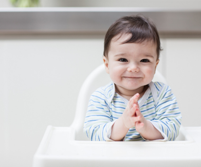 Adorable baby in blue and white striped onesie sitting in a white highchair in a sun filled room.