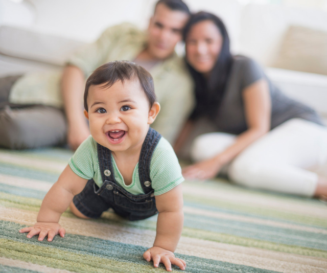 Baby boy crawling across rug with parents laughing in the background.
