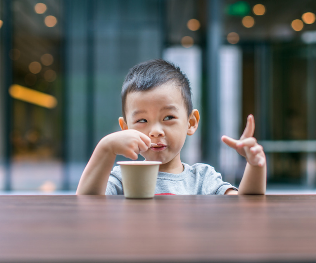 Cheeky toddler with milk moustache sitting with a cup at an outdoor table.
