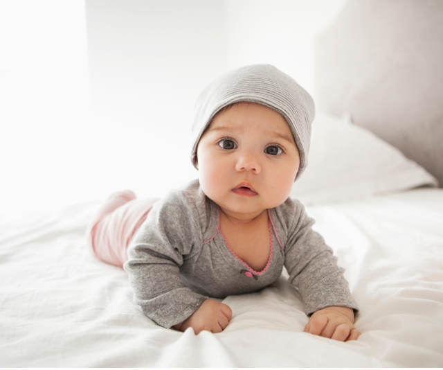 Adorable chubby cheeked baby doing tummy time on bed with white linen while wearing a grey knit onesie and beanie style hat while sun streams in the window.