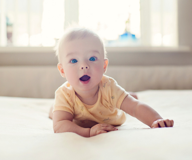 Cute baby in yellow onesie laying on tummy on bed with white linen while sun streams in the window.