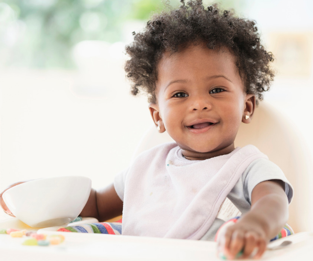 Adorable curly haired baby sitting in high chair, wearing bib and holding a bowl while smiling.