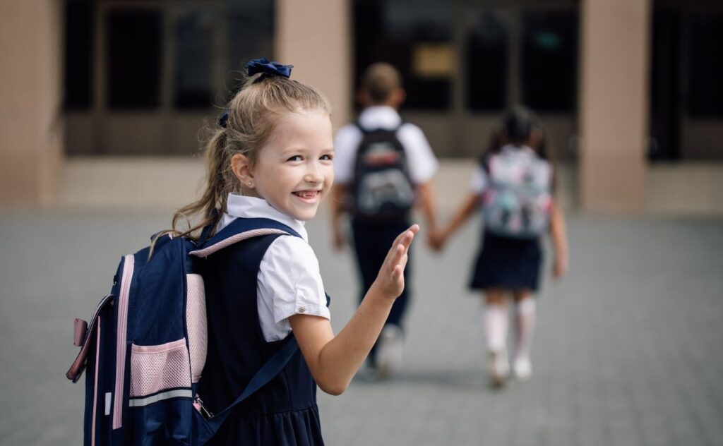 Photo of pre-adolescent blonde girl in school uniform with backpack looking back, smiling and waving to someone behind while walking to school building entrance on first day of school