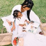 Women and child sit together on picnic blanket on grass, both have long dark braids and are wearing white