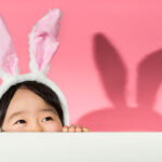 Young dark haired girl wearing bunny ears peering over wall with a pink background.