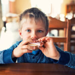 Cute little boy eating breakfast sandwich with ham and cheese in restaurant or home. The boy aged 5 and looking at the camera while biting the sandwich.
