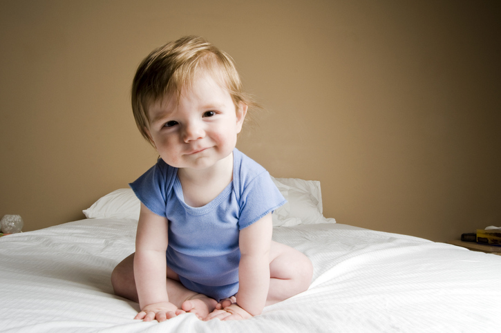 Red headed baby in blue onesie sitting on a bed