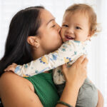 Longhaired woman kissing smiling child's cheek in front of curtained window