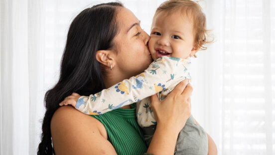 Longhaired woman kissing smiling child's cheek in front of curtained window