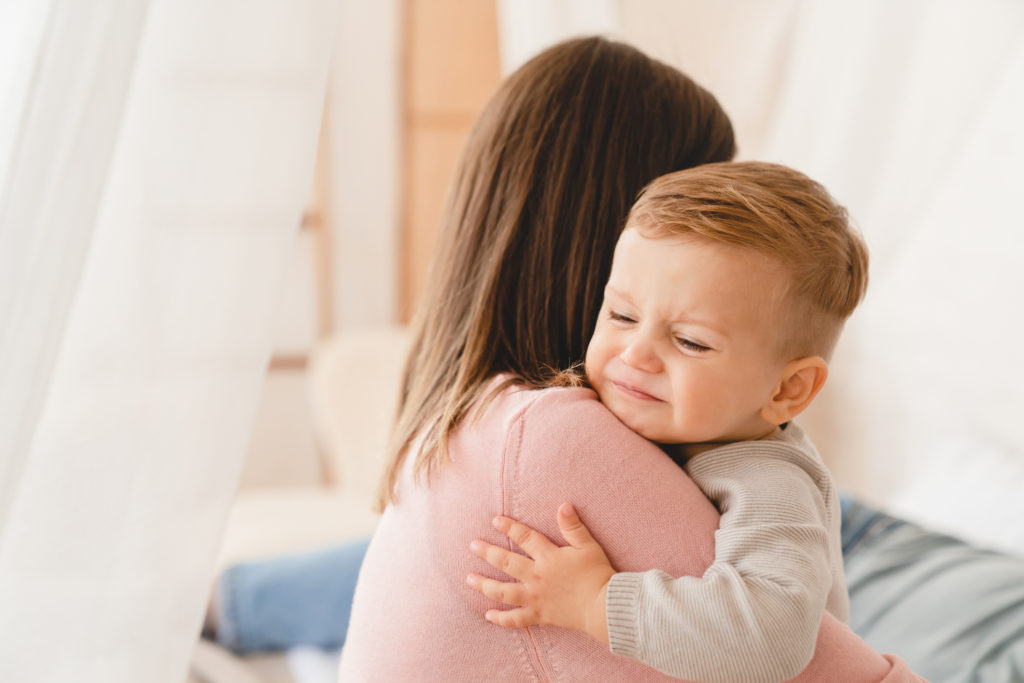 Toddler boy with his eyes closed and a grimace on his face cuddling a woman with long dark hair