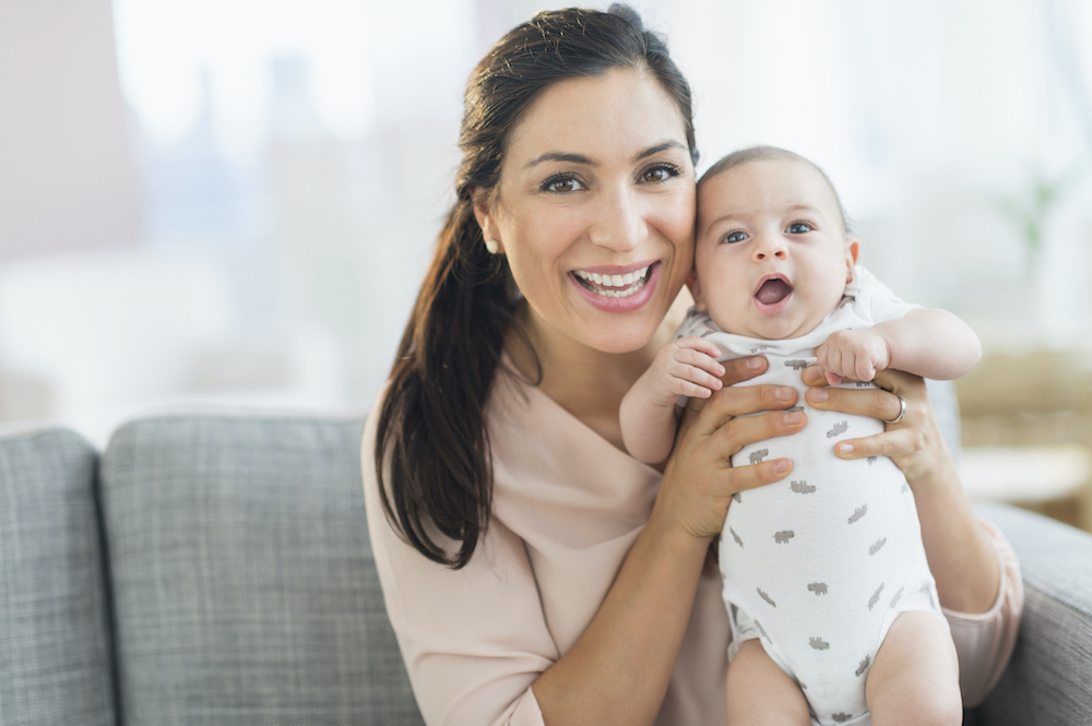 Mum with long dark hair holding her baby under the arms and smiling at the camera