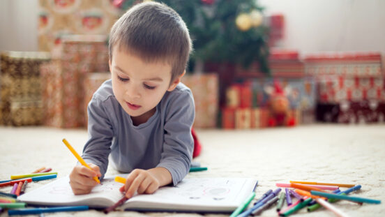 Adorable little boy, writing with colorful pencils in a book, lying on the floor at home, Christmas tree and presents behind him.