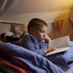 Primary school boy lying on his side reading a book in bed