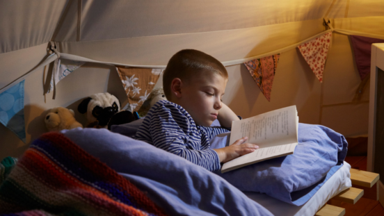 Primary school boy lying on his side reading a book in bed