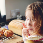 Little girls looking back towards the camera, licking her finger, a tray of hot cross buns nearby