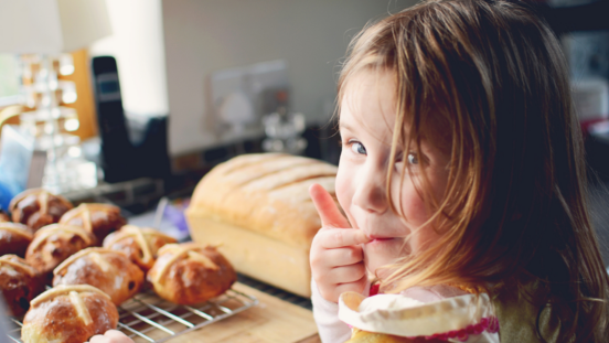 Little girls looking back towards the camera, licking her finger, a tray of hot cross buns nearby