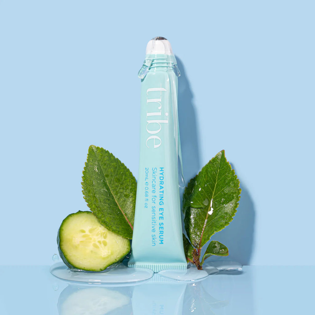 Pale blue bottle of Tribe Hydrating Eye Serum with herb leaves and a cucumber slice