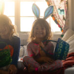 Two cute kids wearing bunny ears sit in bed holding Easter baskets and wearing pajamas, candid happy photo of two kids getting ready to enjoy the holiday