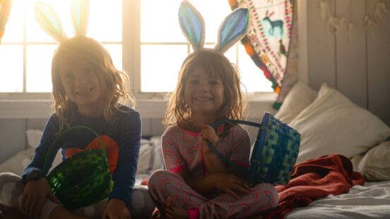 Two cute kids wearing bunny ears sit in bed holding Easter baskets and wearing pajamas, candid happy photo of two kids getting ready to enjoy the holiday