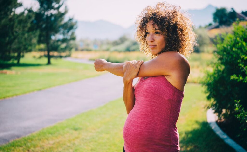 A pregnant woman in a pink singlet stretching and exercising in a park.