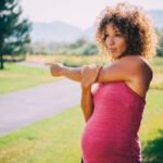A pregnant woman in a pink singlet stretching and exercising in a park.