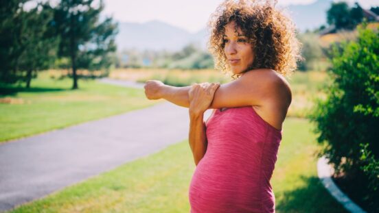 A pregnant woman in a pink singlet stretching and exercising in a park.