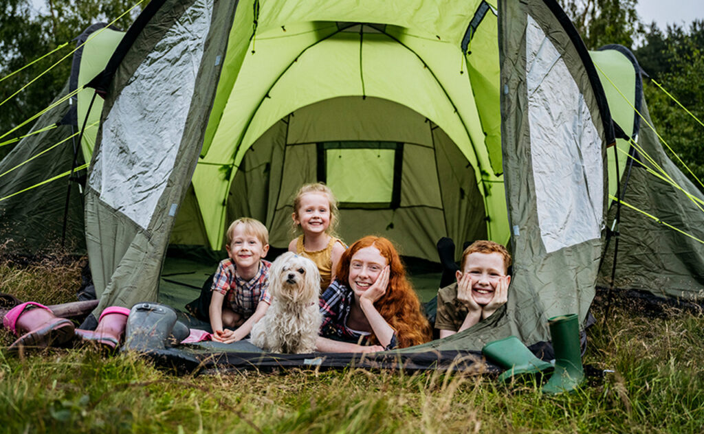 Front view of grinning children aged 5-15 lying on fronts inside tent opening with dog and smiling at camera.