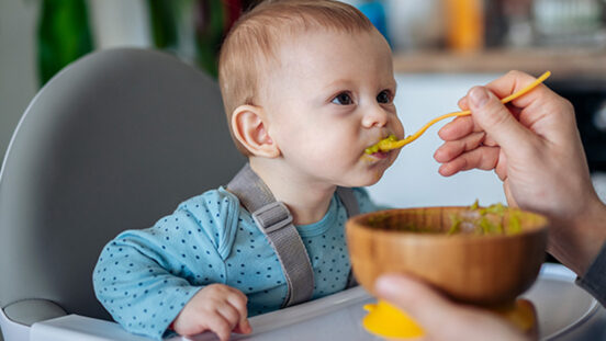 Small brown eyed boy being spoon fed while he sits in a portable highchair