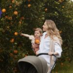 Woman with long blond hair pushing a stroller and carrying a toddler on her hip as they walk by an orange bush