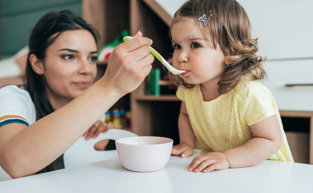 Young, dark-haired mum spoon feeds soup to her cute toddler daughter