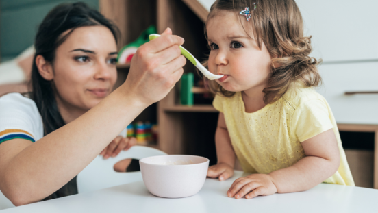 Young, dark-haired mum spoon feeds soup to her cute toddler daughter