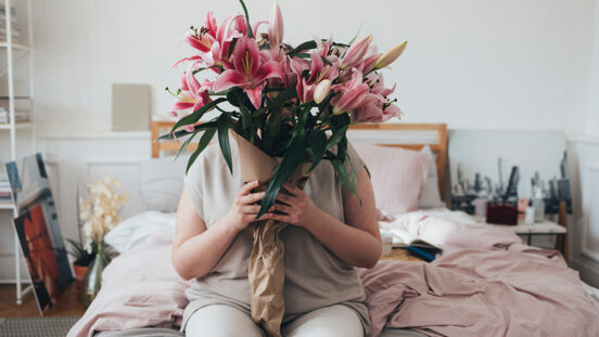 An overweight woman sitting on her bed and holding a bouquet of pink lillies