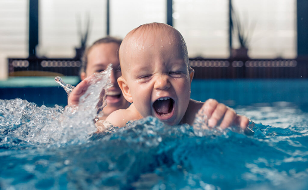 Nine month old baby boy at his first swimming lesson. Looking at camera, smiling and enjoying water. Mother is holding him and helping him to swim.