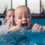 Nine month old baby boy at his first swimming lesson. Looking at camera, smiling and enjoying water. Mother is holding him and helping him to swim.