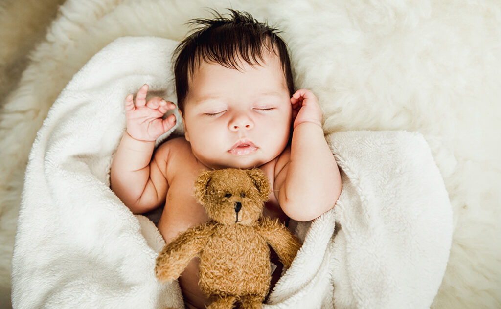 Sleeping dark haired baby wrapped in white blanket with a brown teddy bear nestled on her lap.